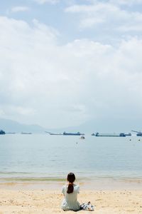 Rear view of woman sitting on beach against cloudy sky
