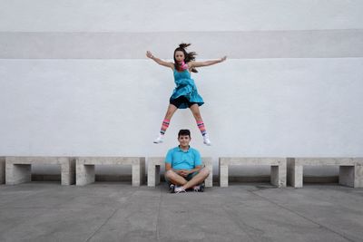 Portrait of young woman jumping over friend sitting against wall
