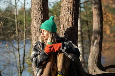 Wistful woman sitting on ground after walking in pine forest, drinking hot tea, warming in sun rays