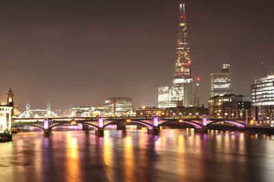 Illuminated bridge over river by buildings against sky at night