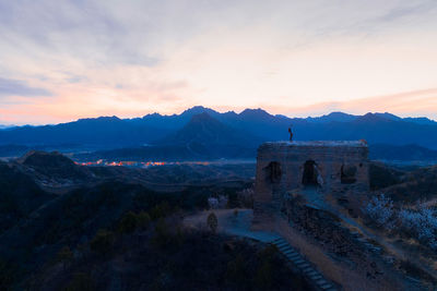 Scenic view of mountains with man standing on built structure against sky during sunset