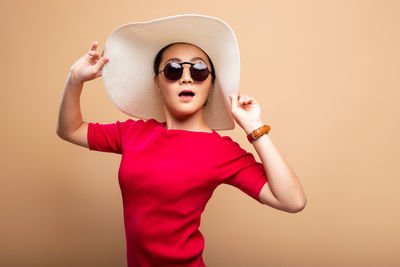 Portrait of woman wearing hat and sunglasses while standing against brown background