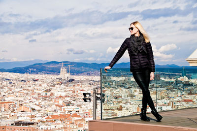 Young woman wearing warm clothing while standing on balcony against cityscape