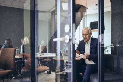 Mature businessman using smart phone seen through glass door at creative office