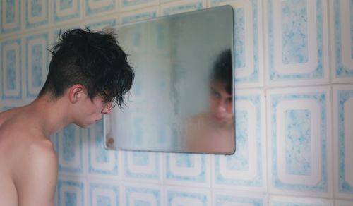 Shirtless man looking at his reflection in mirror at bathroom