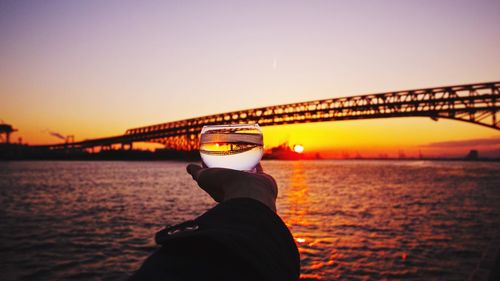 Cropped image of hand holding drinking glass against bridge over river during sunset