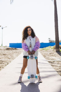 Full body of female with skateboard in hand standing looking at camera on sidewalk along tall palms against coast and sea