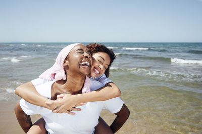 Delighted black mother piggybacking young daughter while having fun with closed eyes at seaside in summer