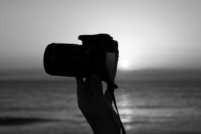 Cropped image of silhouette hand holding camera by sea against sky during sunset