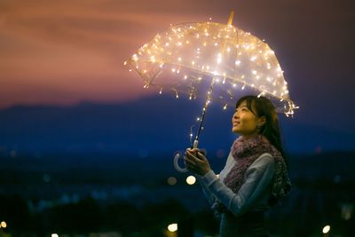 Smiling young woman holding illuminated umbrella against sky at dusk