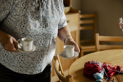 Senior woman holding saucers with tea cups near dining table at home