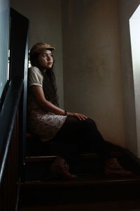 Young woman looking away while sitting on staircase by window at home