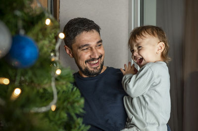 Happy father carrying cute boy in front of christmas tree at home