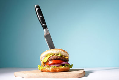 Tasty beef burger on a wooden table with a knife inserted into it on blue background