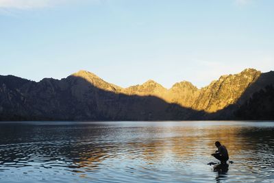 Side view of man crouching amidst lake with mountains in background