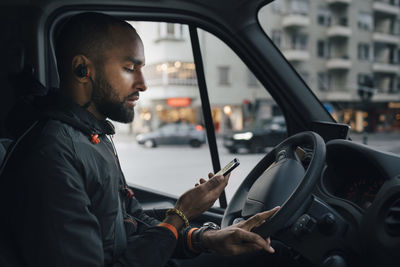 Side view of male driver looking at mobile phone while sitting in delivery van at city