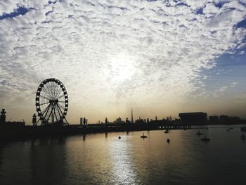 Silhouette of ferris wheel by river against cloudy sky