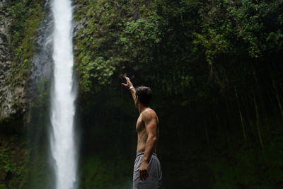 Rear view of shirtless man standing against waterfall in forest