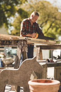 Low angle view of senior man cutting wooden plank with hand saw at yard