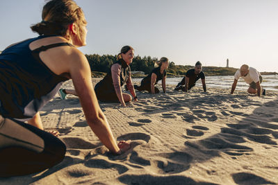 Smiling female instructor doing stretching with team on sand at beach