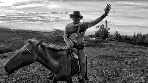 Man gesturing while sitting on horse against cloudy sky