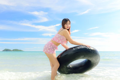 Portrait of beautiful woman holding inflatable raft at beach against sky