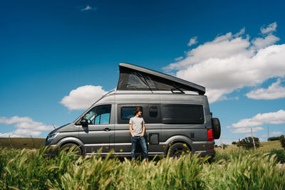 Full body of young male adventurer looking away while resting near camper van with tent parked in summer countryside