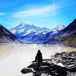 Rear view of mid adult woman sitting on rock by lake against sky during sunny day