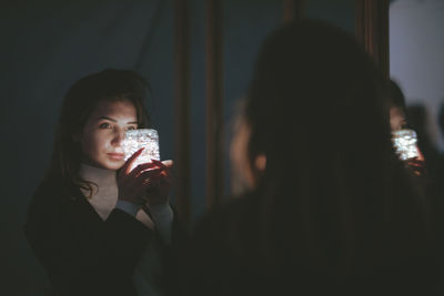 Rear view of young woman holding illuminated lighting equipment reflecting on mirror in darkroom