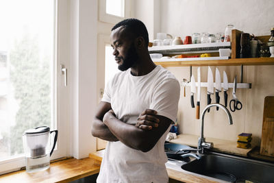 Thoughtful man with arms crossed looking down while leaning on kitchen counter at home