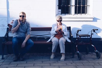 Mother and son sitting on bench by mobility walker against wall during sunny day