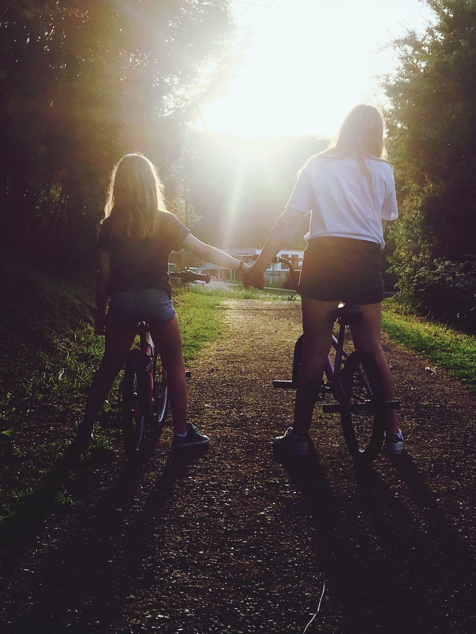 two people, bicycle, togetherness, childhood, full length, leisure activity, bonding, real people, rear view, girls, casual clothing, riding, cycling, day, happiness, outdoors, friendship, lifestyles, tree, sunlight, bmx cycling, grass, cycling helmet, nature, young adult, people