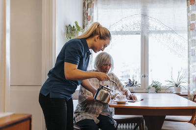 Side view of female caregiver serving tea to senior woman sitting at dining table
