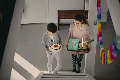 High angle view of siblings carrying birthday cake and gift box while climbing up steps at home