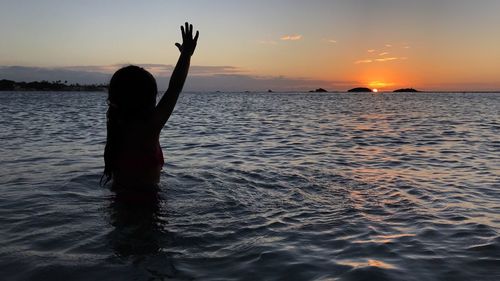 Silhouette girl waving while standing in sea during sunset