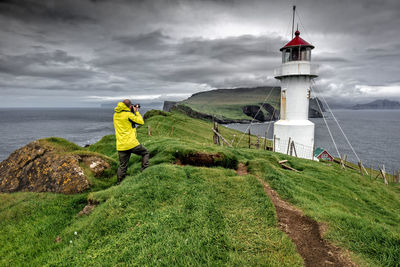 Man photographing lighthouse while standing at beach against cloudy sky