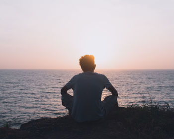 Rear view of man sitting against sea during sunset