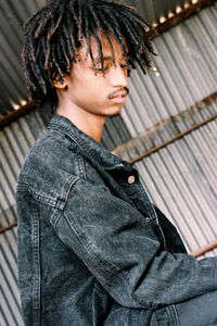 Side view of young man with dreadlocks sitting against corrugated iron