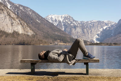 Young man lying outdoors on bench and enjoying mountains, snow, good weather, blue sky, sun. 