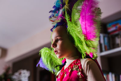 From below serious cute little girl with bright makeup and in colorful a traditional feather headgear carnival costume smiling looking at camera during carnival