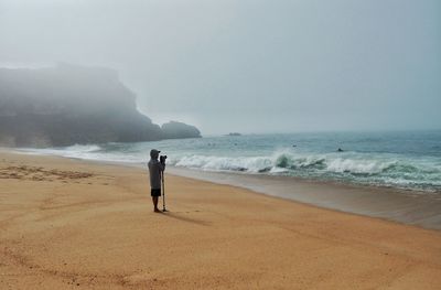 Side view of young man photographing while standing at beach against clear sky