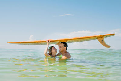 Couple poses underneath surfboard while standing in ocean