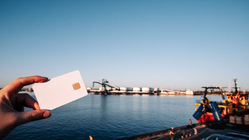 Cropped hand of woman holding paper boat in sea against clear blue sky