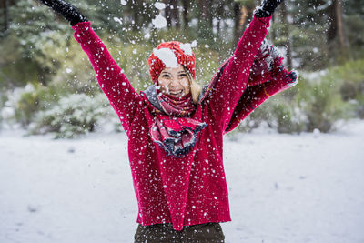 Cheerful young woman throwing snow mid air while standing on snow covered land