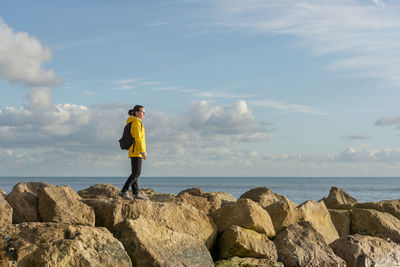 Woman wearing a yellow coat and backpack walking across rocks by the sea