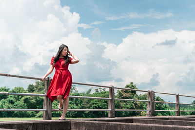 Woman wearing red dress standing by railing against sky