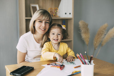 Portrait of a smiling mother and daughter who draw with colored pencils at the table