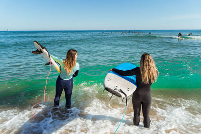 Back view of unrecognizable young female surfers in wetsuits standing with surfboards on the sea getting ready to catch waves in summer day