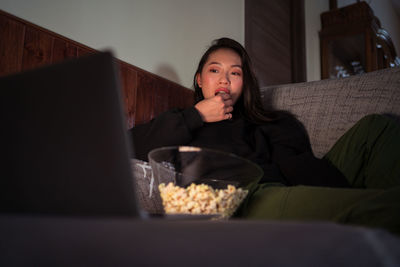 Young asian female wearing casual clothes sitting on couch and eating popcorn at home watching movie on laptop