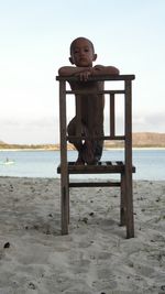 Portrait of boy sitting on chair at beach against sky